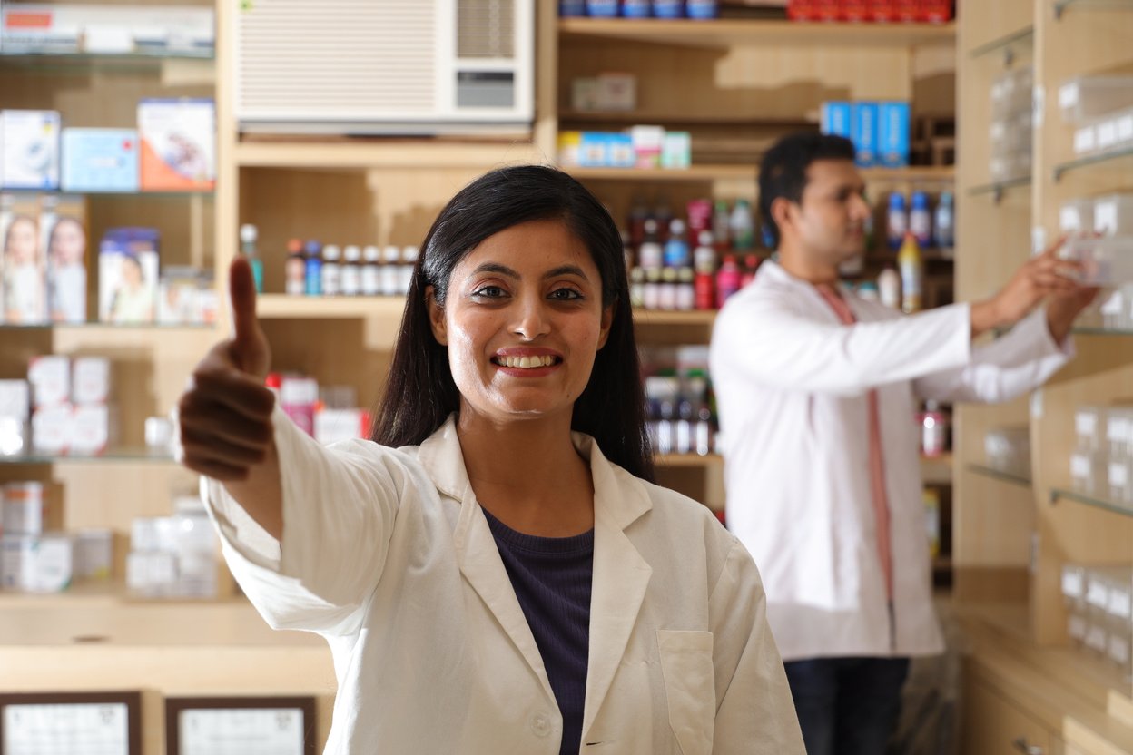 Pharmacist chemist woman standing in pharmacy - drugstore. Close up of pharmacist at the hospital pharmacy. Pharmacist assisting to customers in pharmacy. Showcasing business card in hand.